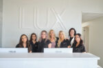 A group of seven women pose behind a desk in front of the word LUX.