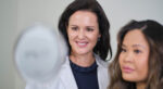 A woman looks at herself in a hand-held mirror while a woman in a lab coat also looks on.