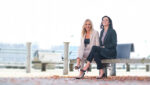 Two women sit on a bench outside, facing the camera and smiling.
